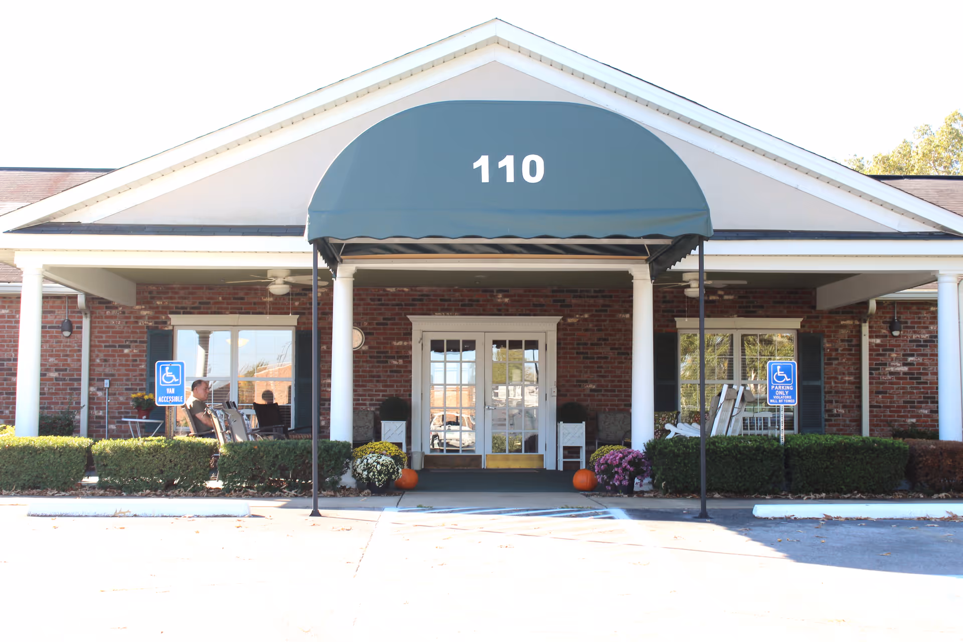 Front entrance of Olive Branch Senior Living facility with a green awning displaying the number 110. The entrance has double glass doors and is flanked by white columns. There are two handicap parking signs visible near the entrance, along with some bushes, flowers, and pumpkins decorating the area. A person is sitting on a chair on the left side porch.