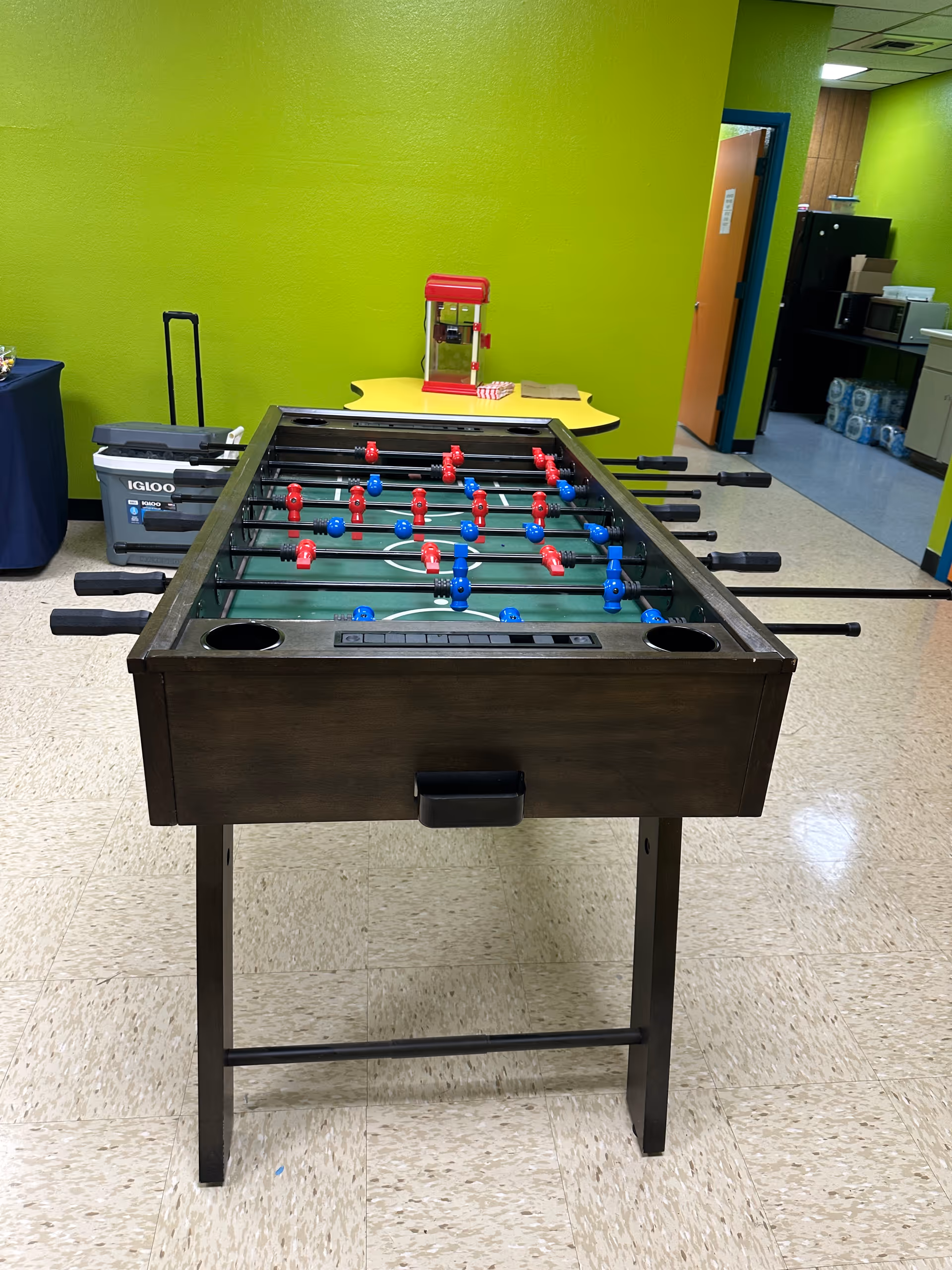 Indoor recreation area with a foosball table, bright green walls, and a hallway in the background.