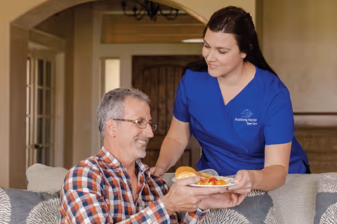 Caregiver in blue scrubs serves a plate of food to an elderly man indoors.