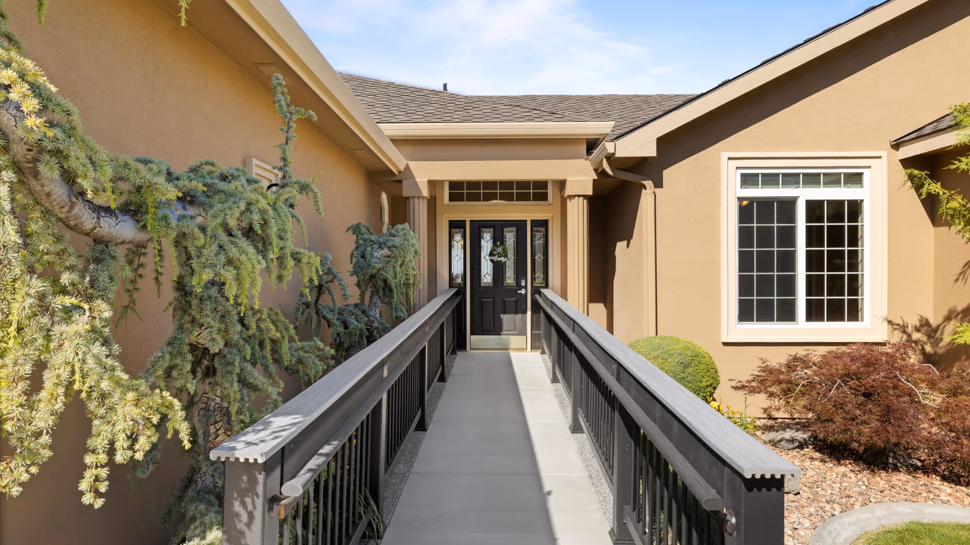 Beige senior living facility entrance with a ramp and black railings leading to double doors, a window to the right, and landscaped shrubs.