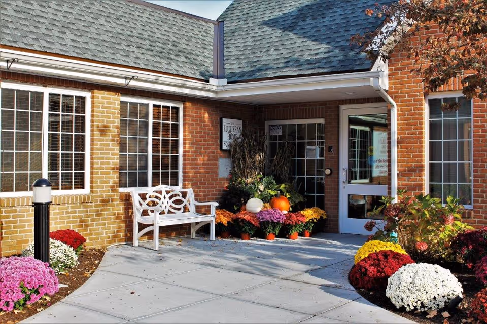 Brick senior living facility entrance with colorful flowers, pumpkins, and a white decorative bench.