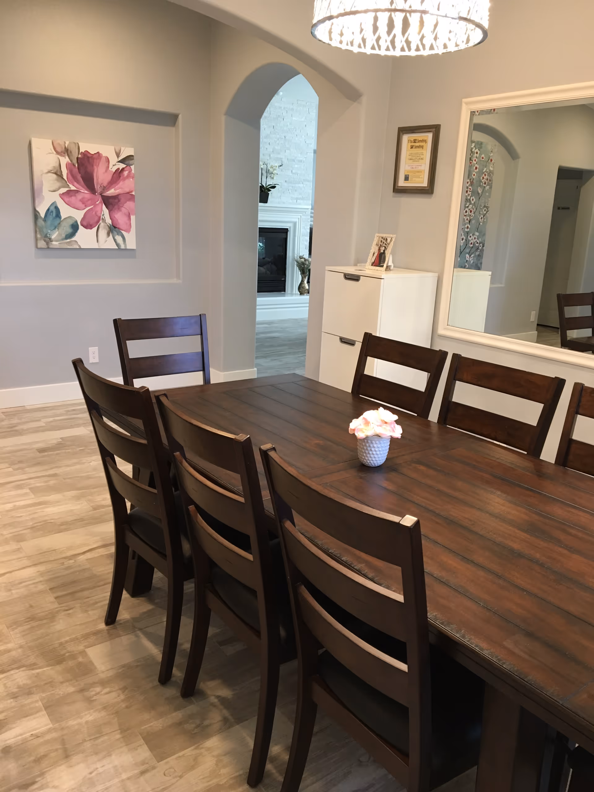 A dining room with a long wooden table surrounded by eight matching wooden chairs. A small vase with pink flowers is placed in the center of the table. The room has light-colored walls, a large mirror on one wall, a white cabinet, and a colorful floral painting. An arched doorway leads to another room with a fireplace.