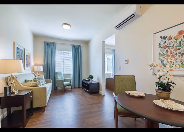 Bright senior living suite with sofa, chairs, dining table, and plants.