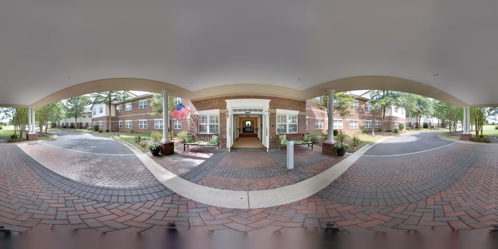 Covered entrance area of a senior living facility with brick-paved driveway and walkways, two benches, potted plants, and an American flag. The building is two stories with brick and white siding, surrounded by trees and greenery.