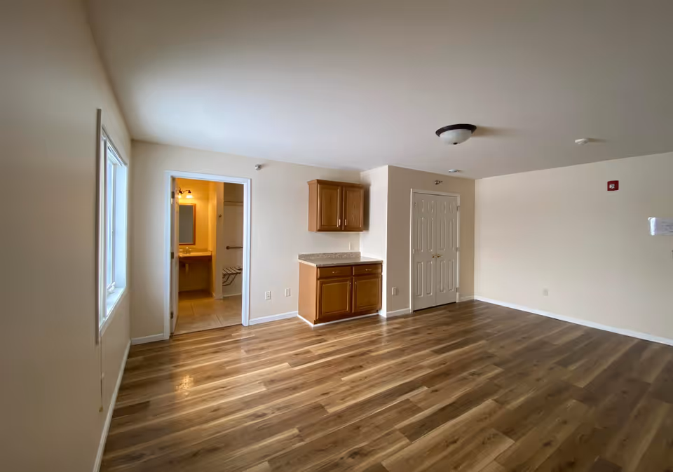 Empty senior living unit with wood floors, small kitchenette, and bathroom doorway.