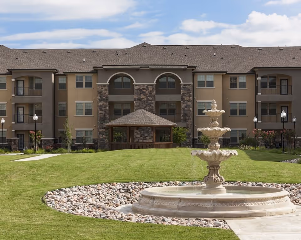 A three-story residential building with balconies and a central gazebo viewed from a lawn with a decorative fountain in the foreground.