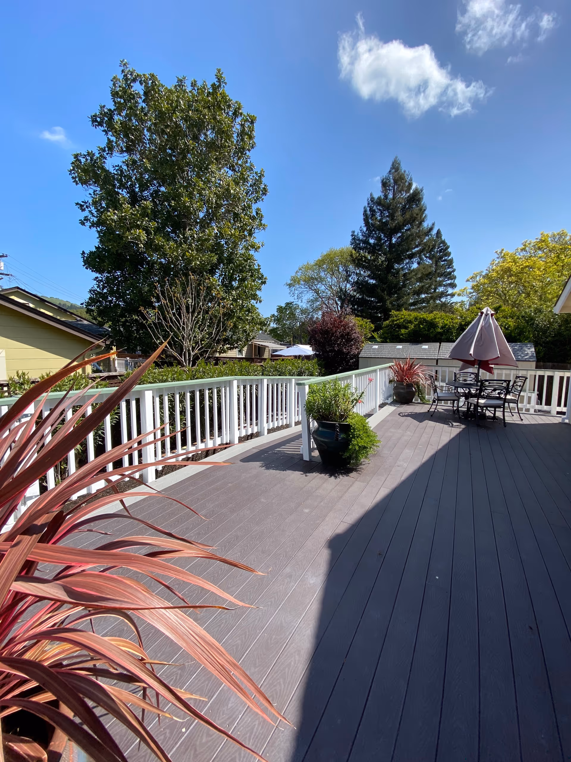 Outdoor deck area with gray wooden flooring, white railing, potted plants, and a patio table with chairs and an umbrella under a clear blue sky with some clouds.