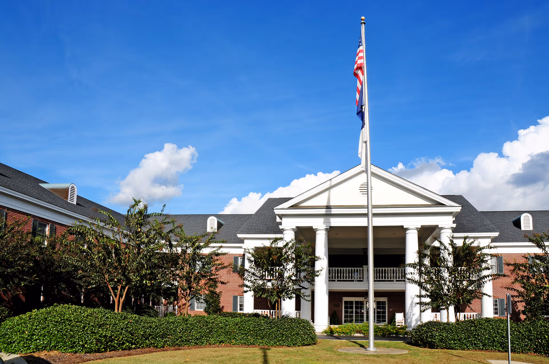 Front exterior of a brick building with white columns, landscaping, and an American flag on a flagpole.