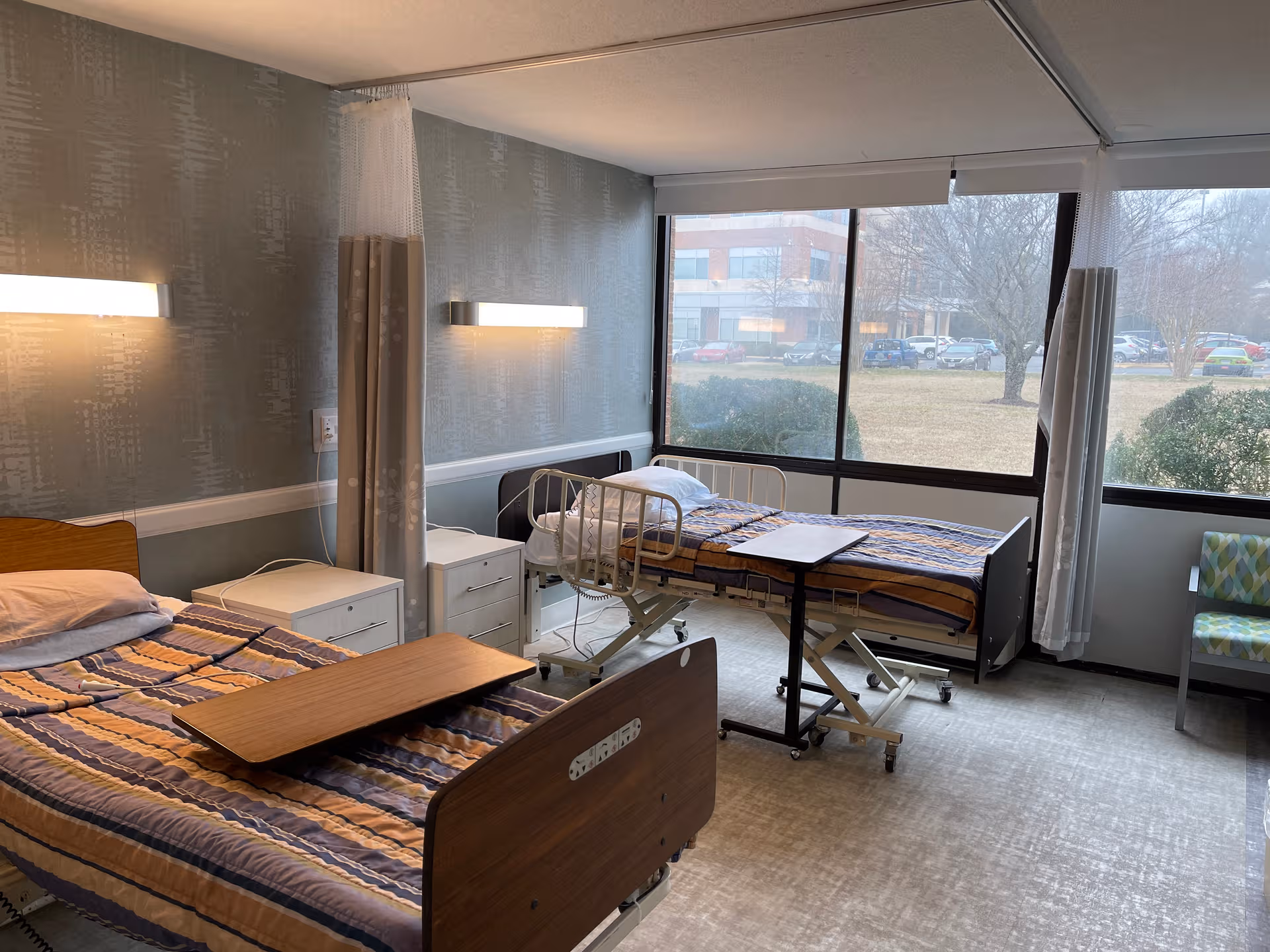 Bright patient room with two hospital beds, nightstands, and large windows.