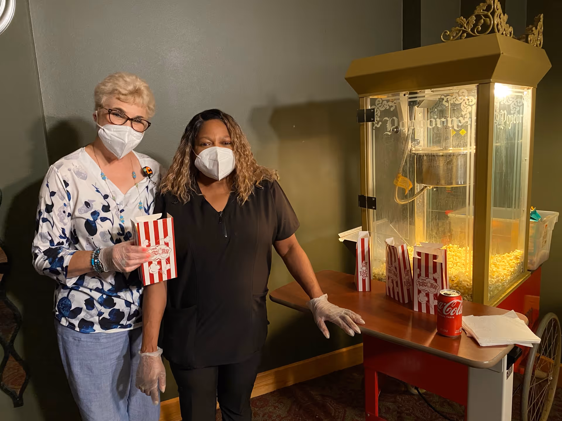 Two women wearing face masks and gloves stand next to a popcorn machine. One woman is holding a striped popcorn bag, and there are several popcorn bags and a can of Coca-Cola on the table beside the machine.