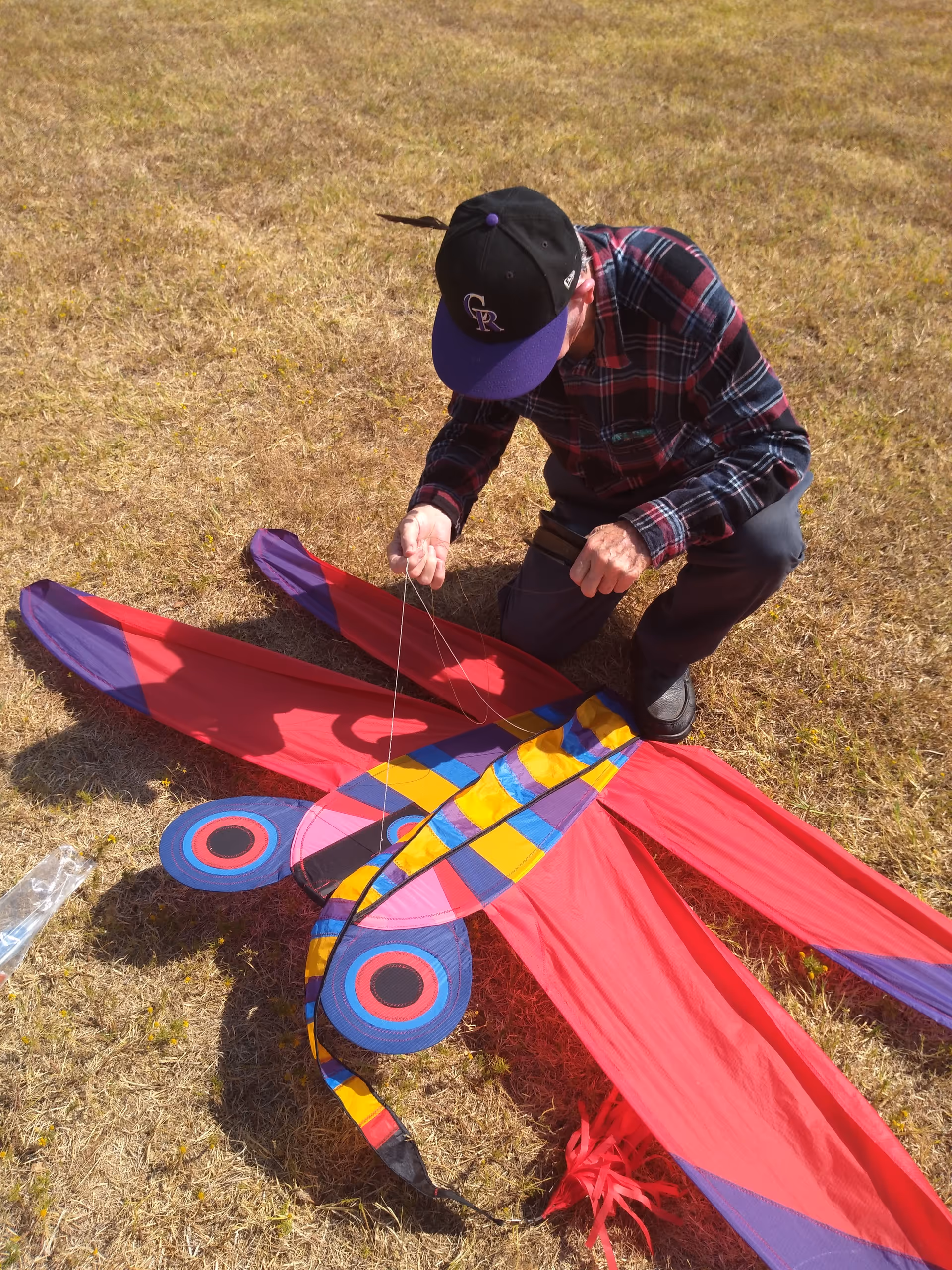 A person kneels on dry grass, assembling a colorful large kite outdoors.