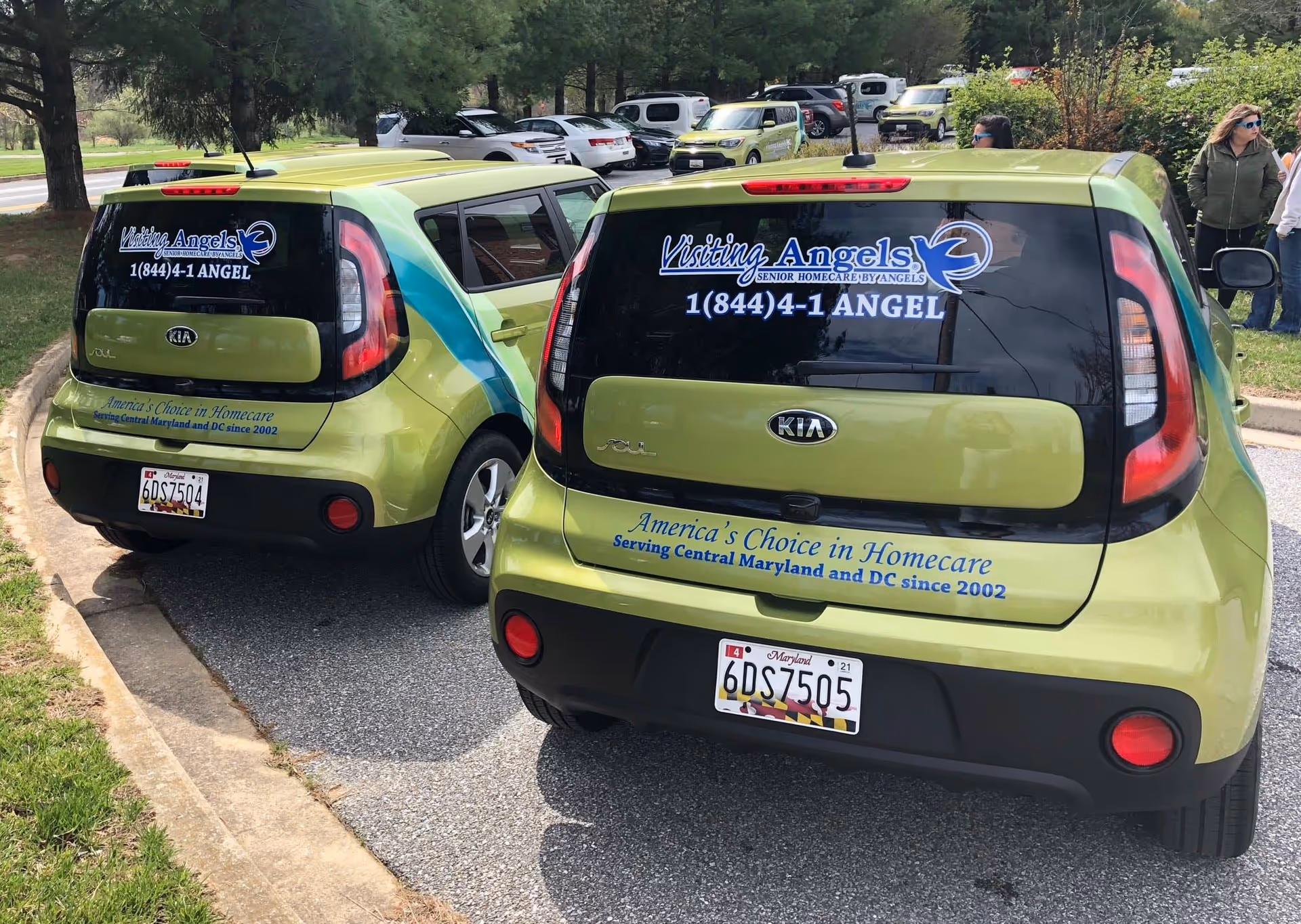 Two lime colored Visiting Angels shuttle vans parked in a lot with people in the background.