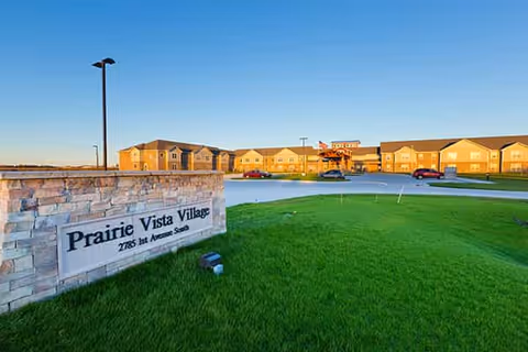 Exterior view of Prairie Vista Village with a stone entrance sign in the foreground, green lawn, parking lot, and the apartment building under a clear blue sky.
