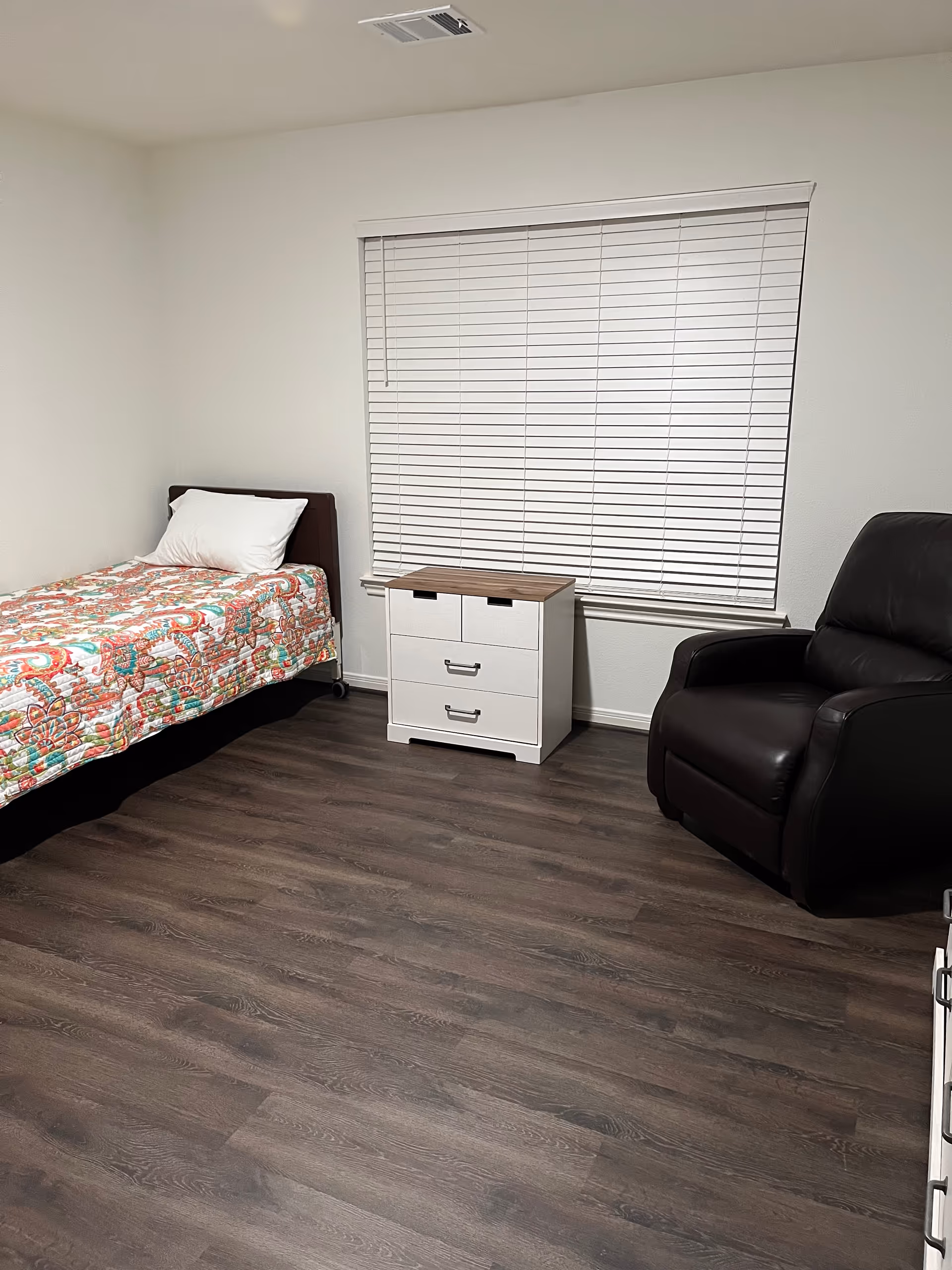 A tidy single bedroom with a bed, white nightstand, blinds, and a recliner.