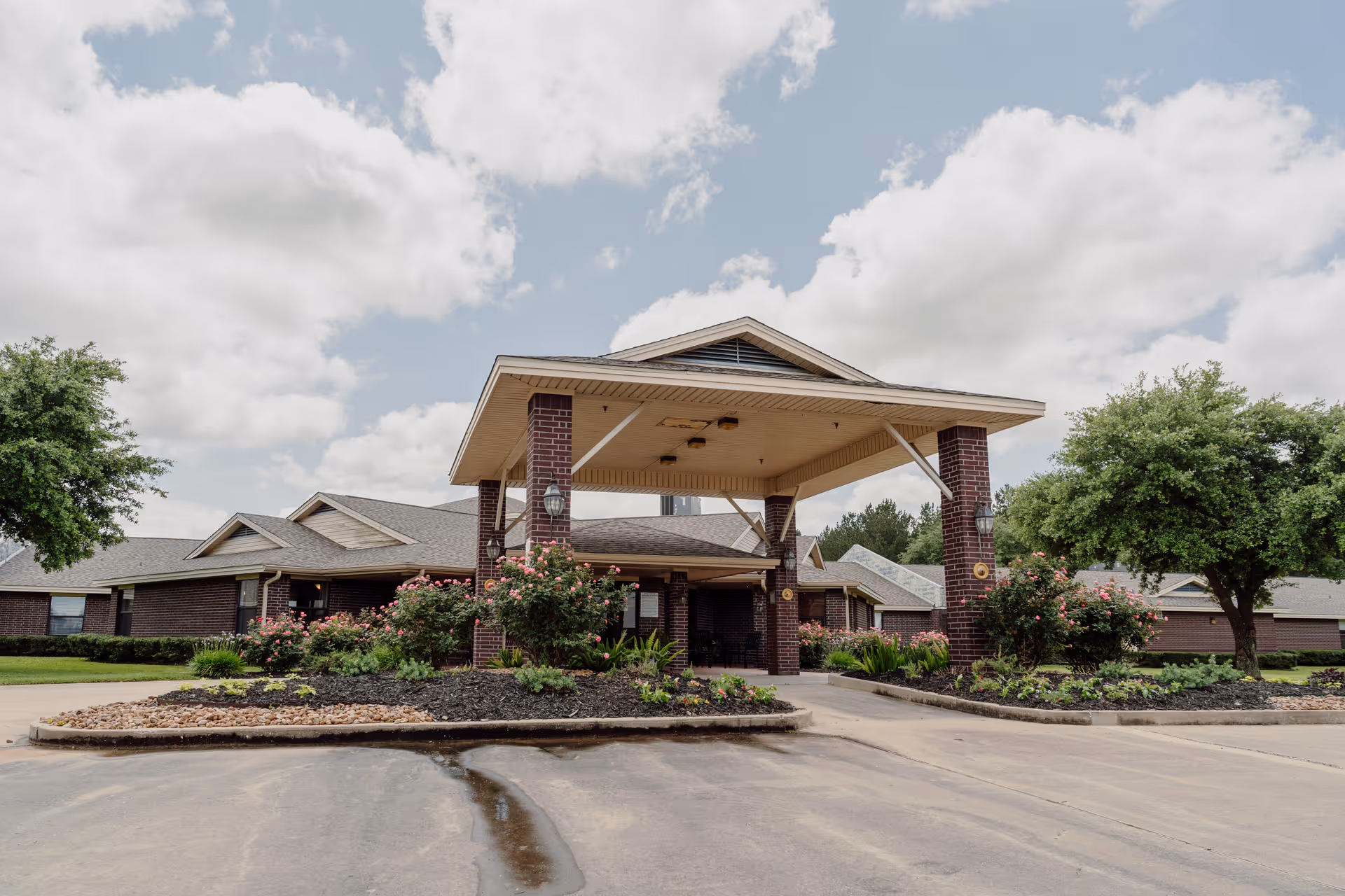 Front exterior view of Pine Ridge Health Care, LLP building with a covered entrance supported by brick pillars, landscaped flower beds, and trees under a partly cloudy sky.