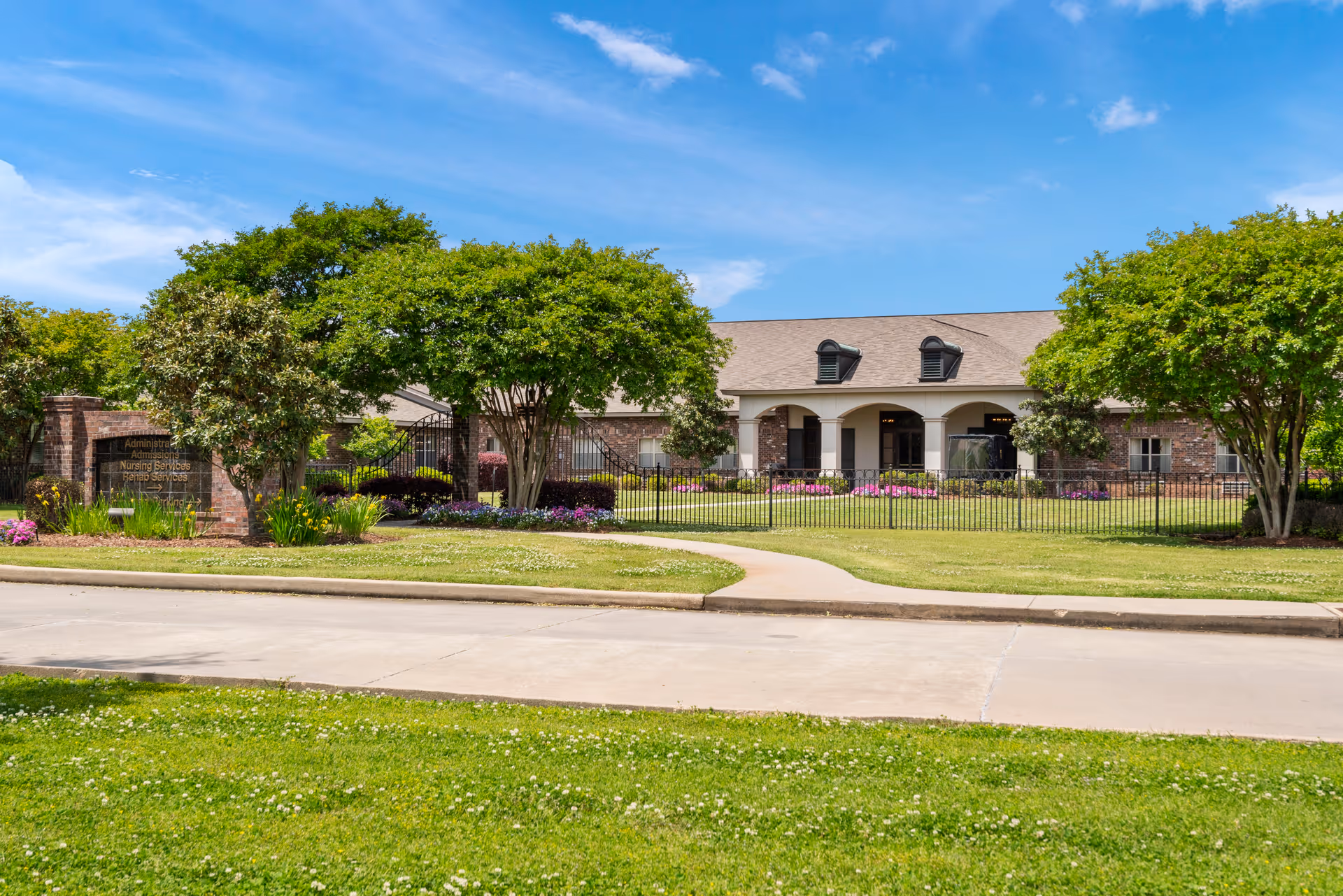 Front exterior of a single-story brick senior living building with a fenced lawn, trees, and a walkway under a blue sky.