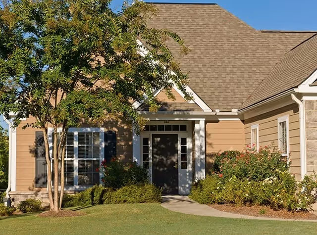 Exterior view of a single-story residential building with beige siding, a brown shingled roof, white trim around the windows and door, surrounded by green shrubs and a tree on a well-maintained lawn under a clear blue sky.