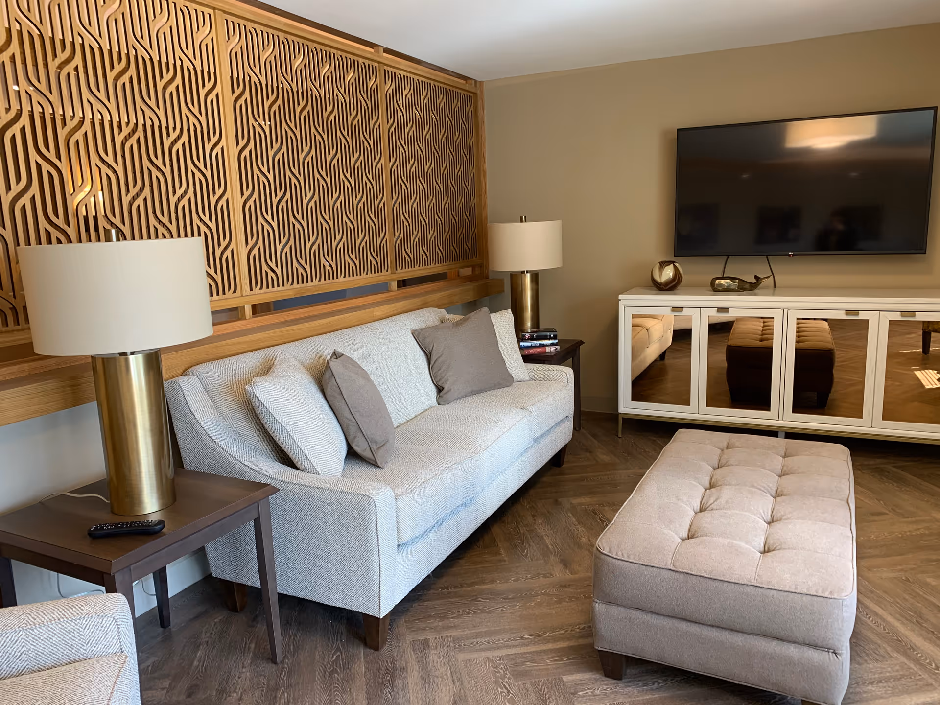Cozy living room with a gray sofa, tufted ottoman, wall-mounted TV above a mirrored console, decorative wood paneling and brass table lamps.