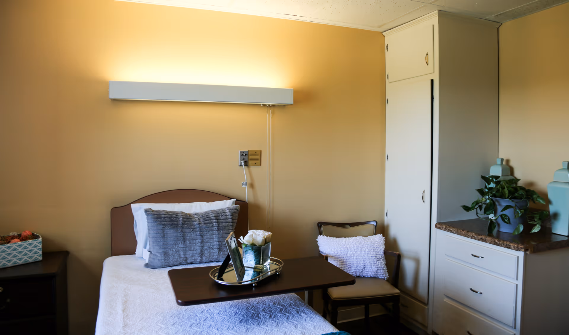 Senior living bedroom with a bed, dresser, chair, and potted plants.
