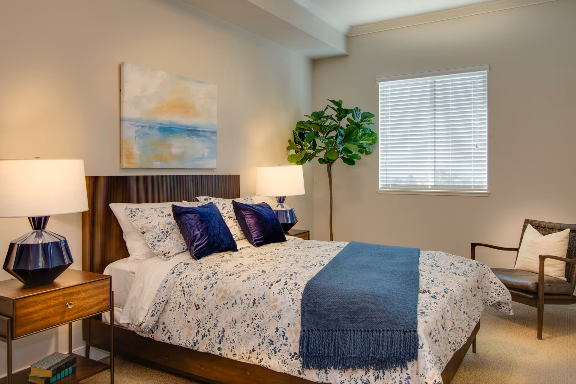 A neatly arranged senior living facility bedroom with a bed, lamps, artwork, and plant.