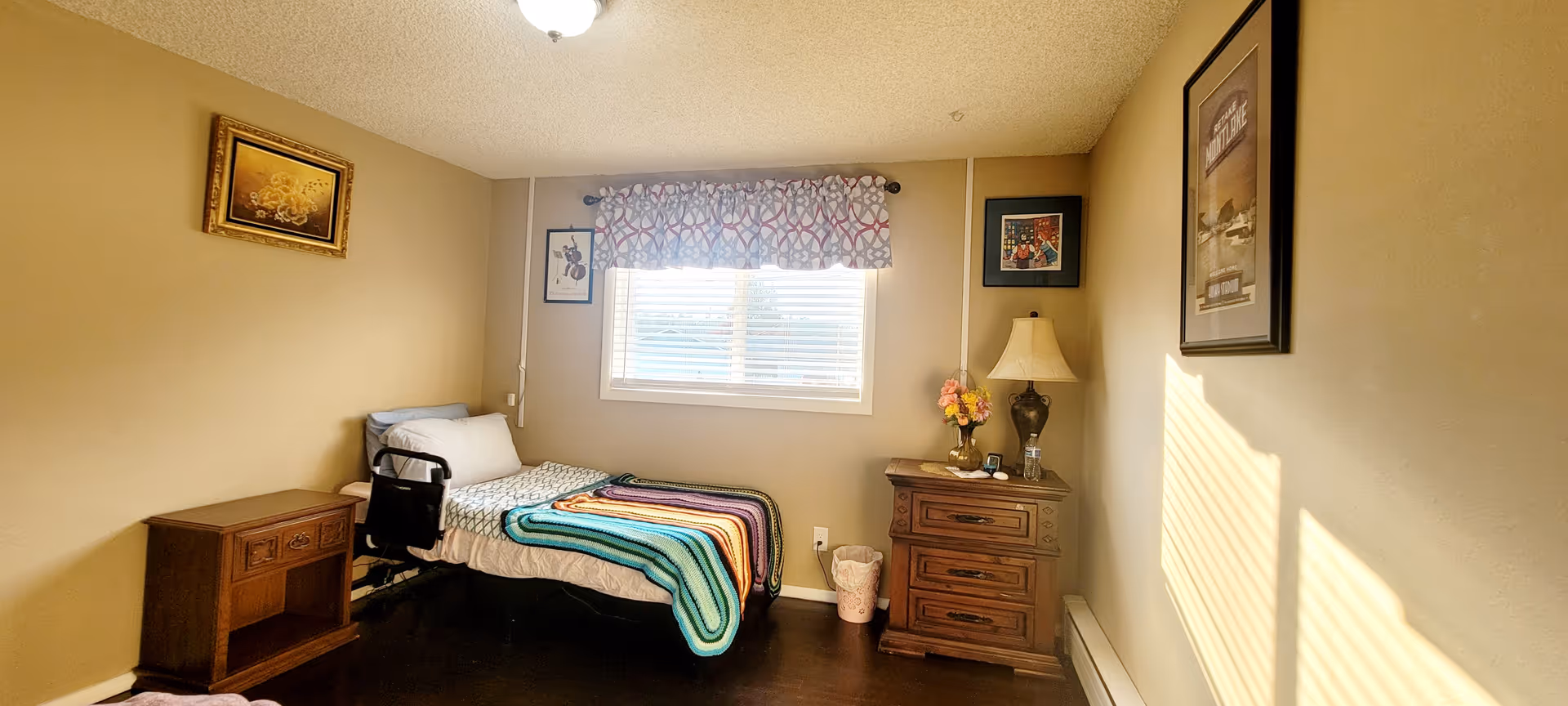A small furnished bedroom with a bed, dresser, window, and wall art.
