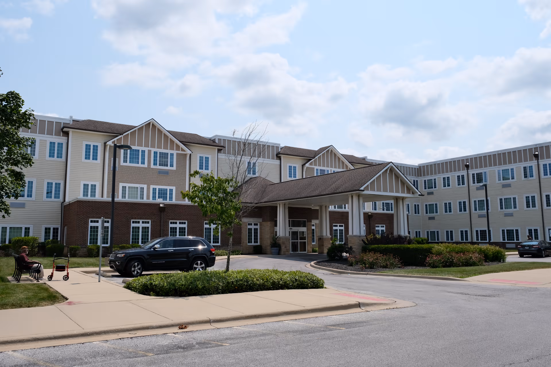 Front exterior of a multi-story senior living building with a covered entrance, driveway, parked cars, and a person sitting on a bench.