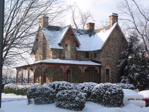 Snow-covered stone building with a pitched roof and a porch, framed by leafless trees.