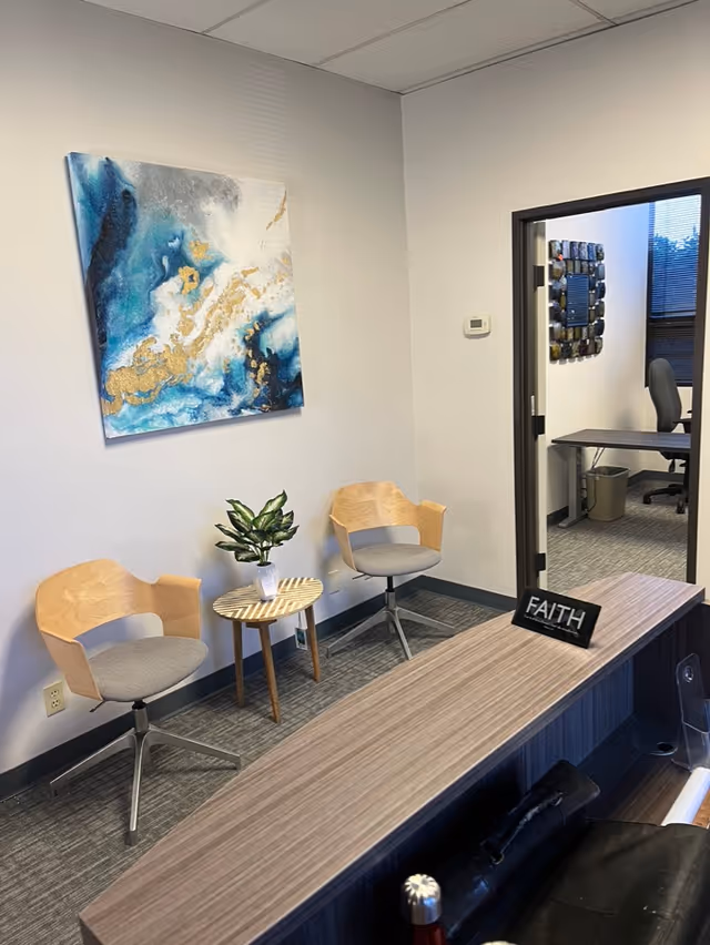 Reception area with two wooden chairs, a small table, plant, abstract artwork, and desk.