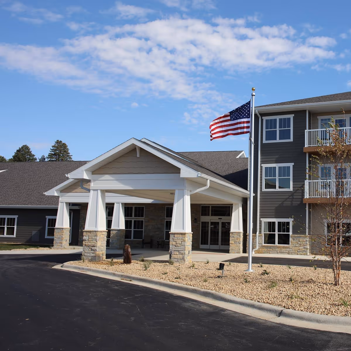 Exterior view of a senior living facility named Winn Prairie featuring a covered entrance with stone pillars, an American flag on a flagpole, and a three-story building with balconies and multiple windows under a partly cloudy blue sky.