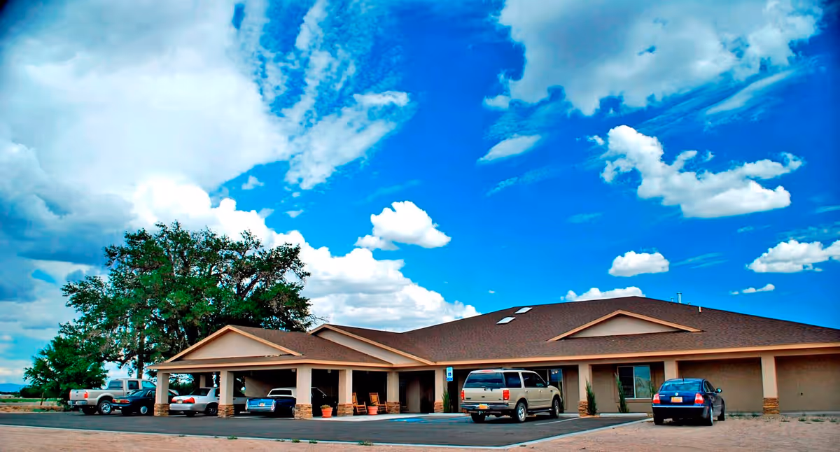 Exterior view of a single-story building with a brown roof and beige walls under a bright blue sky with scattered white clouds. Several cars are parked in front of the building, which has a covered entrance supported by columns. Large trees are visible to the left of the building.