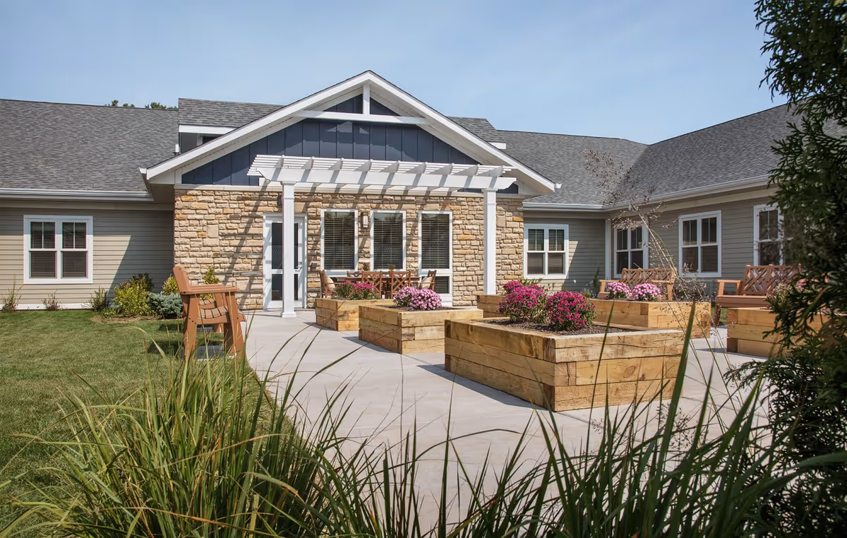 Sunlit courtyard with raised wooden planters, benches, a pergola and the facility's stone-and-siding facade.