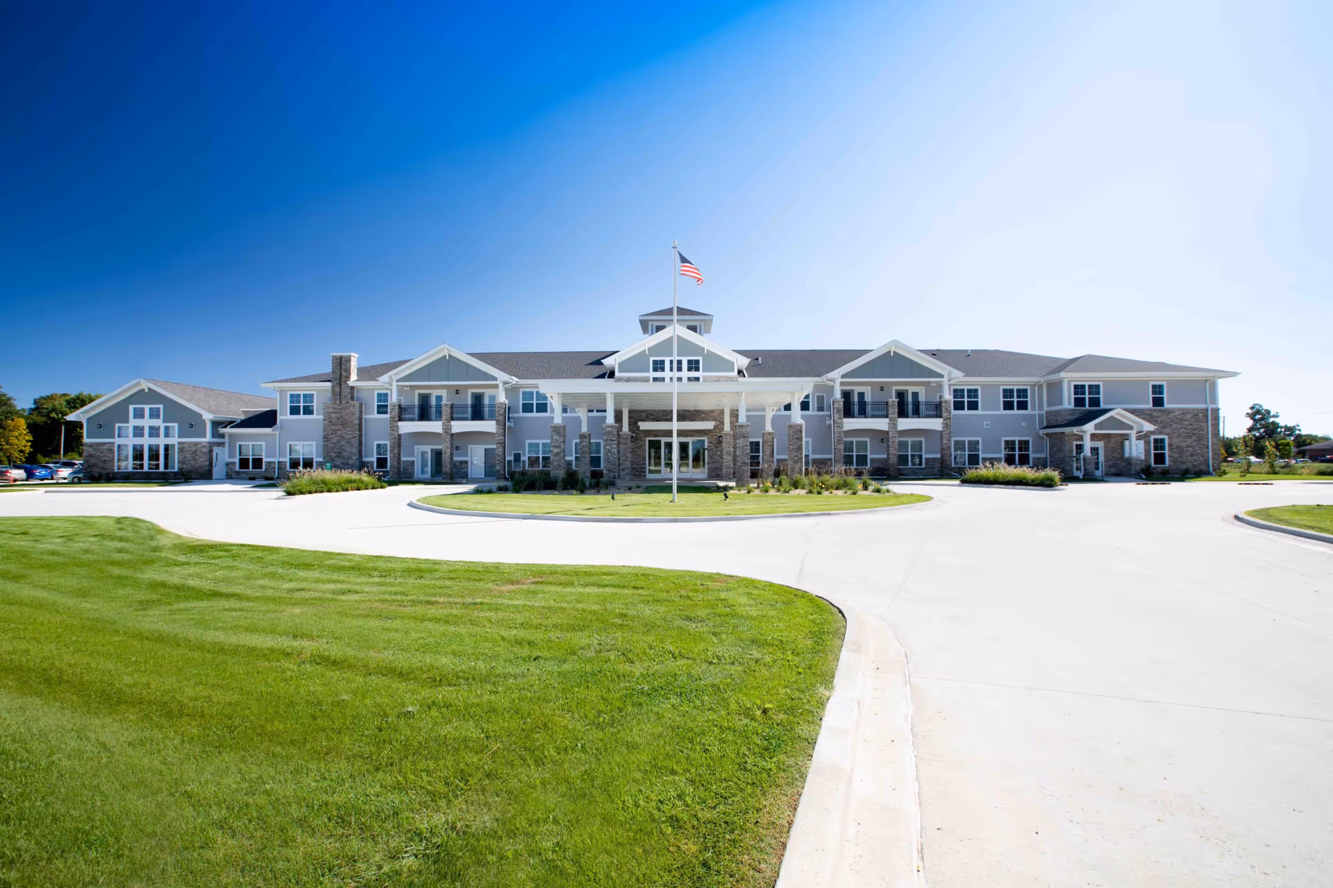 Front exterior of a two-story senior living building with a circular driveway, flagpole, and green lawn under a clear blue sky.