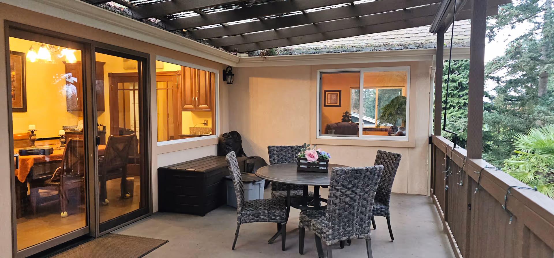 Outdoor balcony with round table, wicker chairs, string lights, and interior kitchen through glass doors.