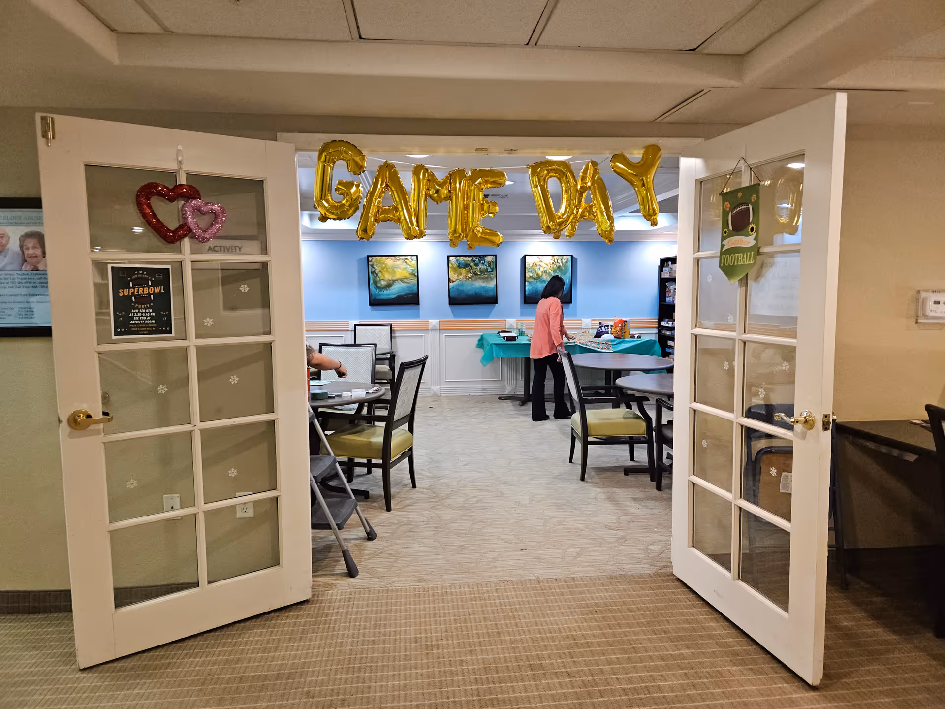 Open French doors reveal a communal activity/dining room decorated with gold 'GAME DAY' balloons, tables and chairs, and a person at a table.