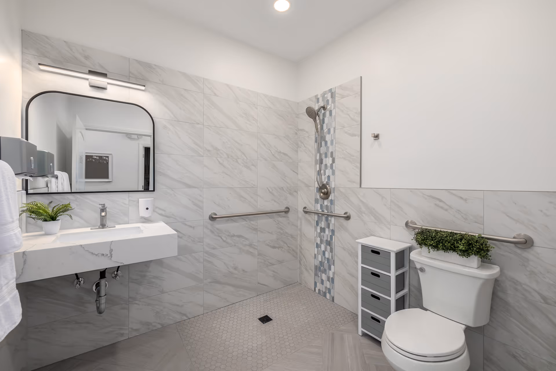 Accessible bathroom with sink, mirror, shower, toilet, and wall grab bars in a tiled space.
