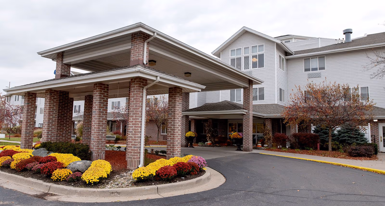 Front entrance of a multi-story senior living building with a covered porte-cochere and colorful flower beds.