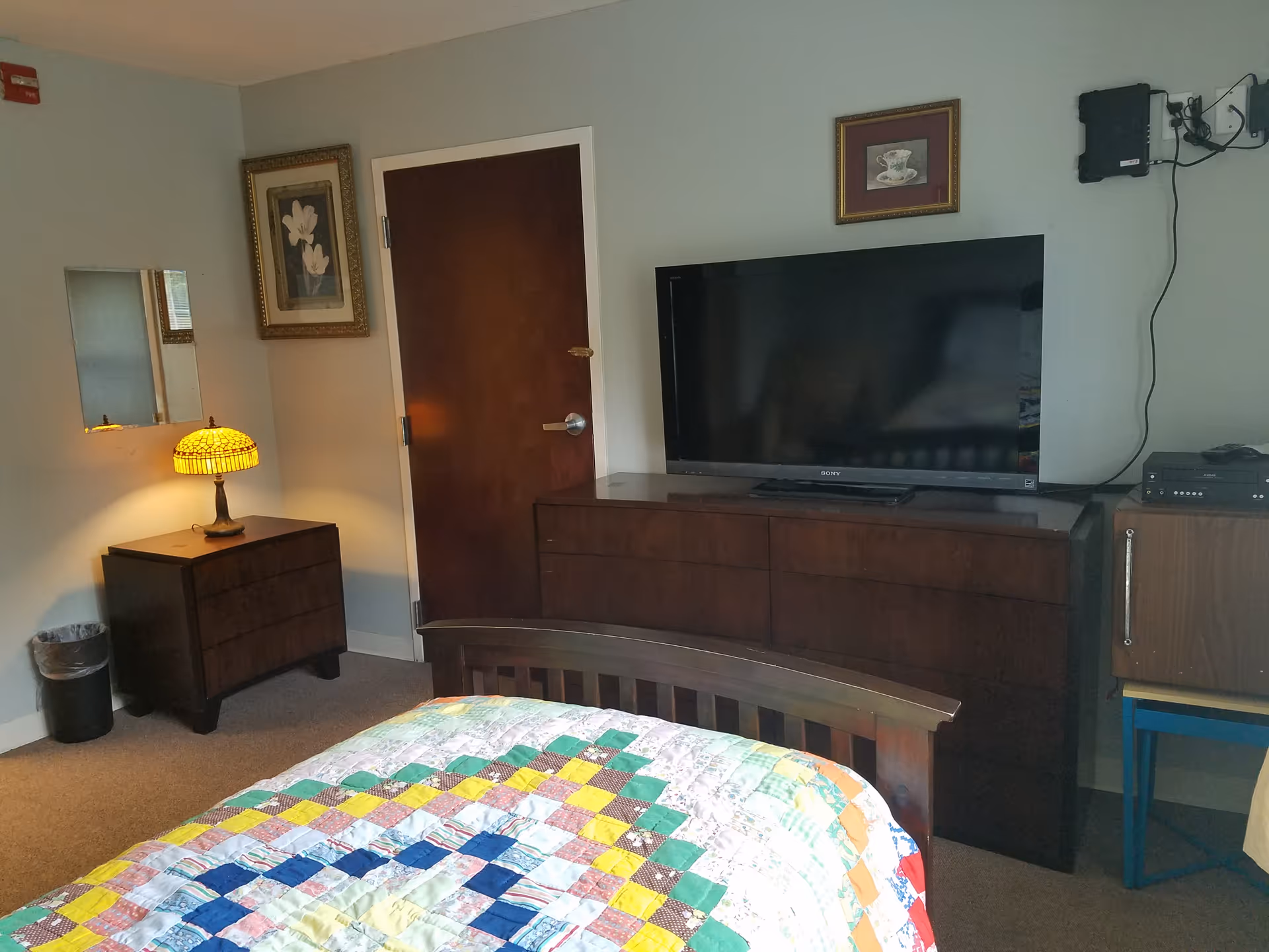 Bedroom in a senior home with a quilted bedspread, dresser, TV, and lamp.
