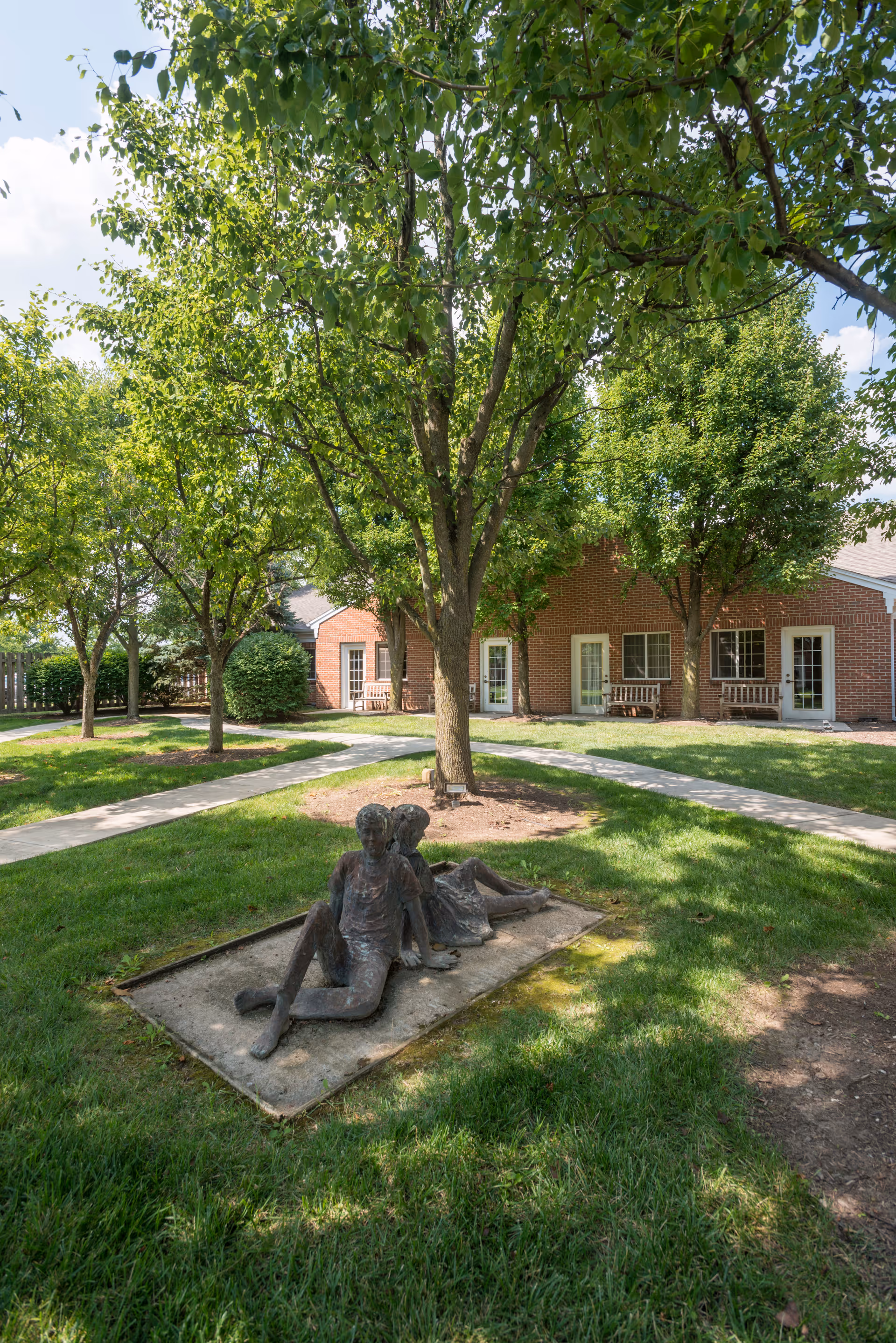 Outdoor garden area with green grass, several trees, and a bronze sculpture of two children sitting back-to-back on a concrete base. In the background, there is a brick building with multiple doors and windows, and wooden benches placed outside.