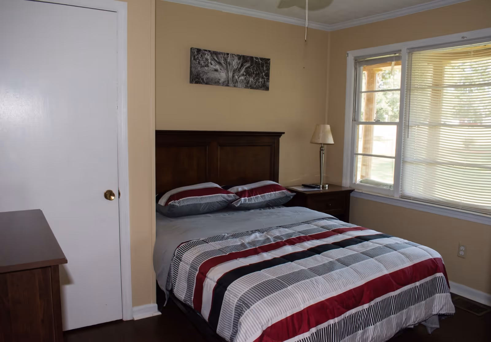 A bedroom with a bed that has a striped comforter in shades of red, gray, black, and white. There are two matching pillows on the bed. To the right of the bed is a wooden nightstand with a lamp and a remote control. A window with blinds is on the right wall, and a white door is on the left side of the room. A black and white picture hangs above the bed on the beige wall.