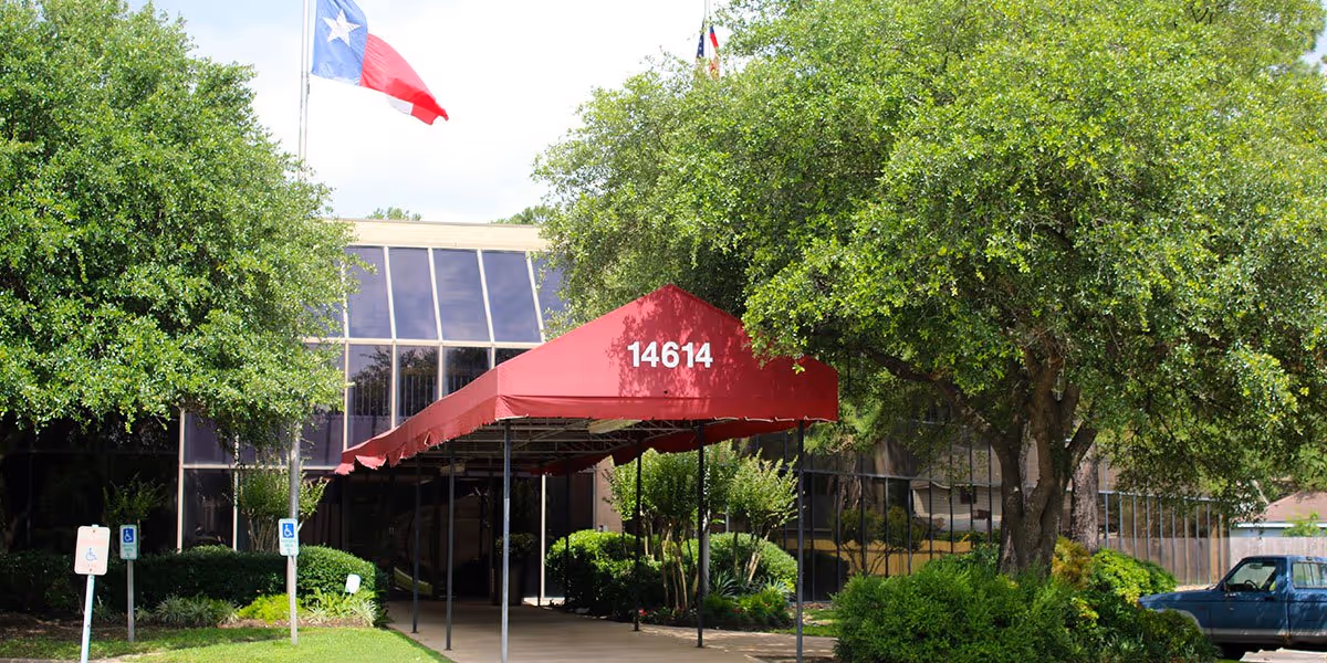 Front entrance of a senior home facility with a red canopy, greenery, and a Texas flag.