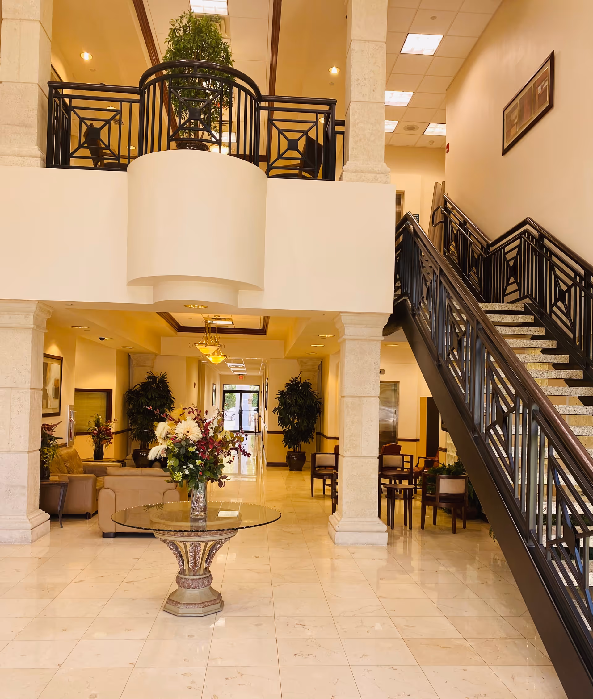Grand hotel-style lobby with a circular balcony, staircase, plants, and a glass-topped round table.
