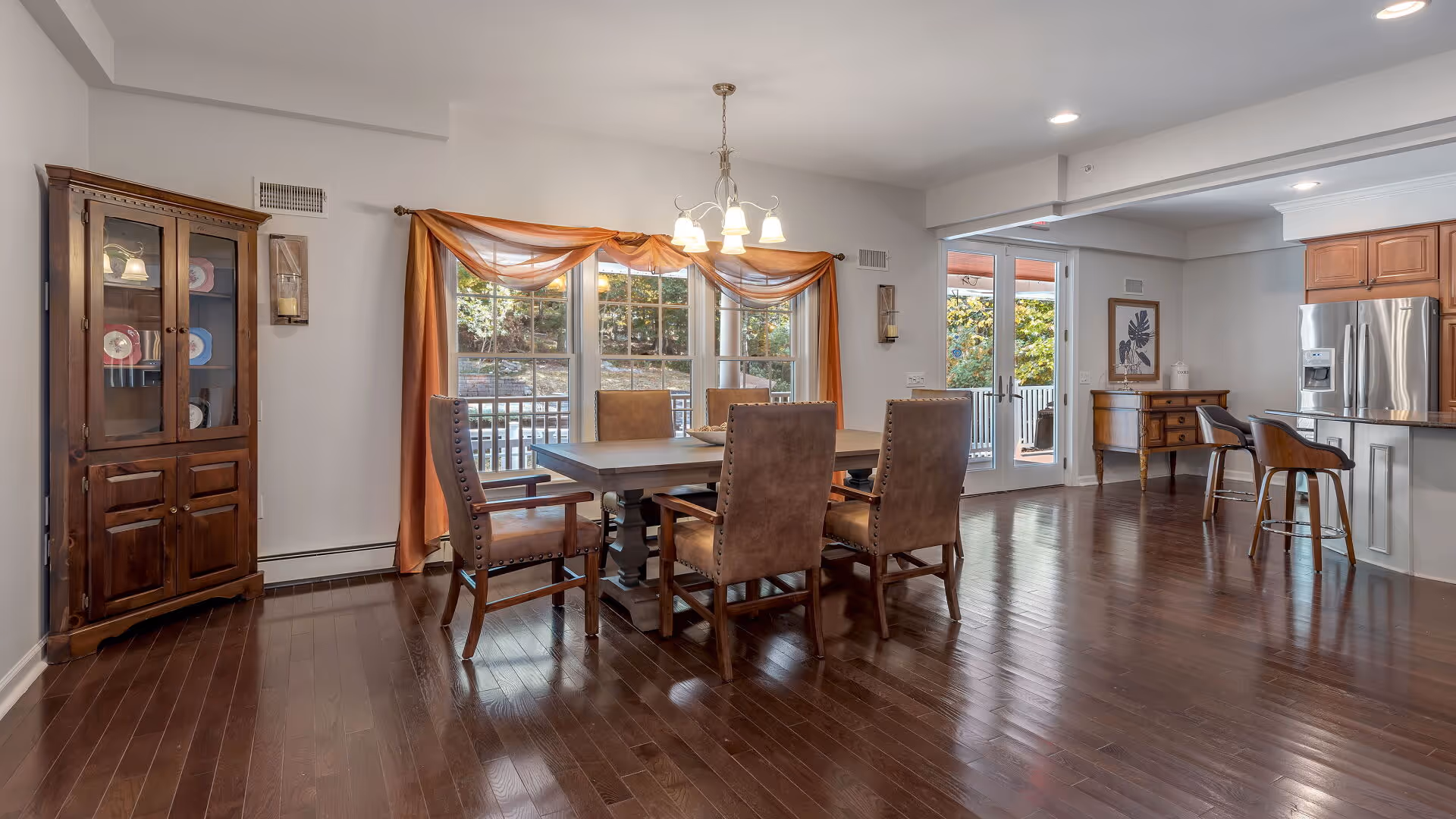 Bright open dining area with a wooden table and upholstered chairs, hardwood floors, a china cabinet, and an adjacent kitchen with a stainless steel refrigerator.