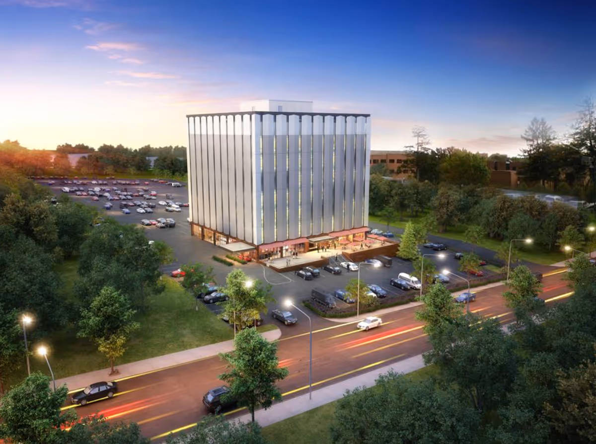 A tall glass-front building with a modern design, parking lot, and surrounding trees.