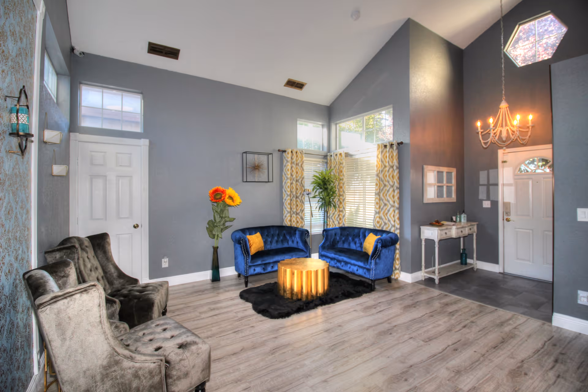 A bright and spacious living room with high ceilings and large windows. The room features two blue velvet loveseats with yellow accent pillows, two gray armchairs, a round gold coffee table on a black rug, and a tall vase with sunflowers. There is a small white console table near the entrance door, a chandelier hanging from the ceiling, and patterned yellow and white curtains on the windows.
