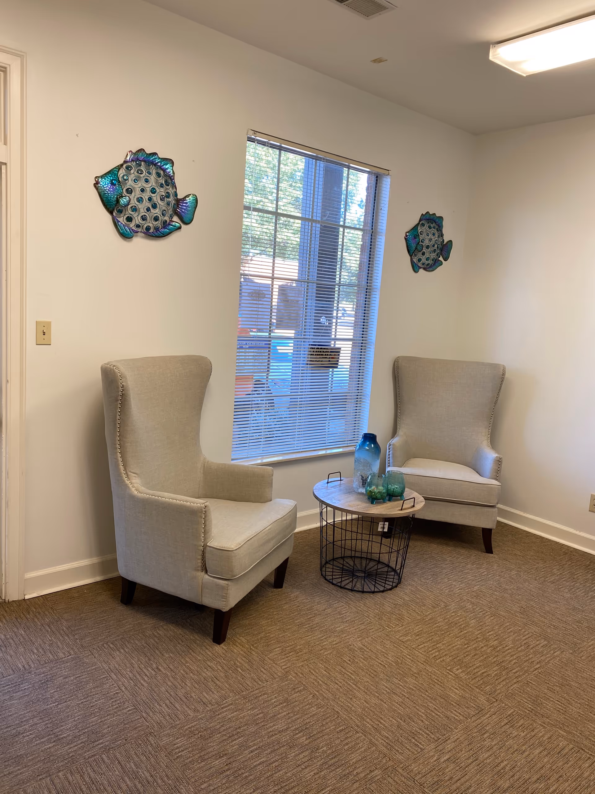 Two beige armchairs flank a round coffee table in a small sunlit living room.