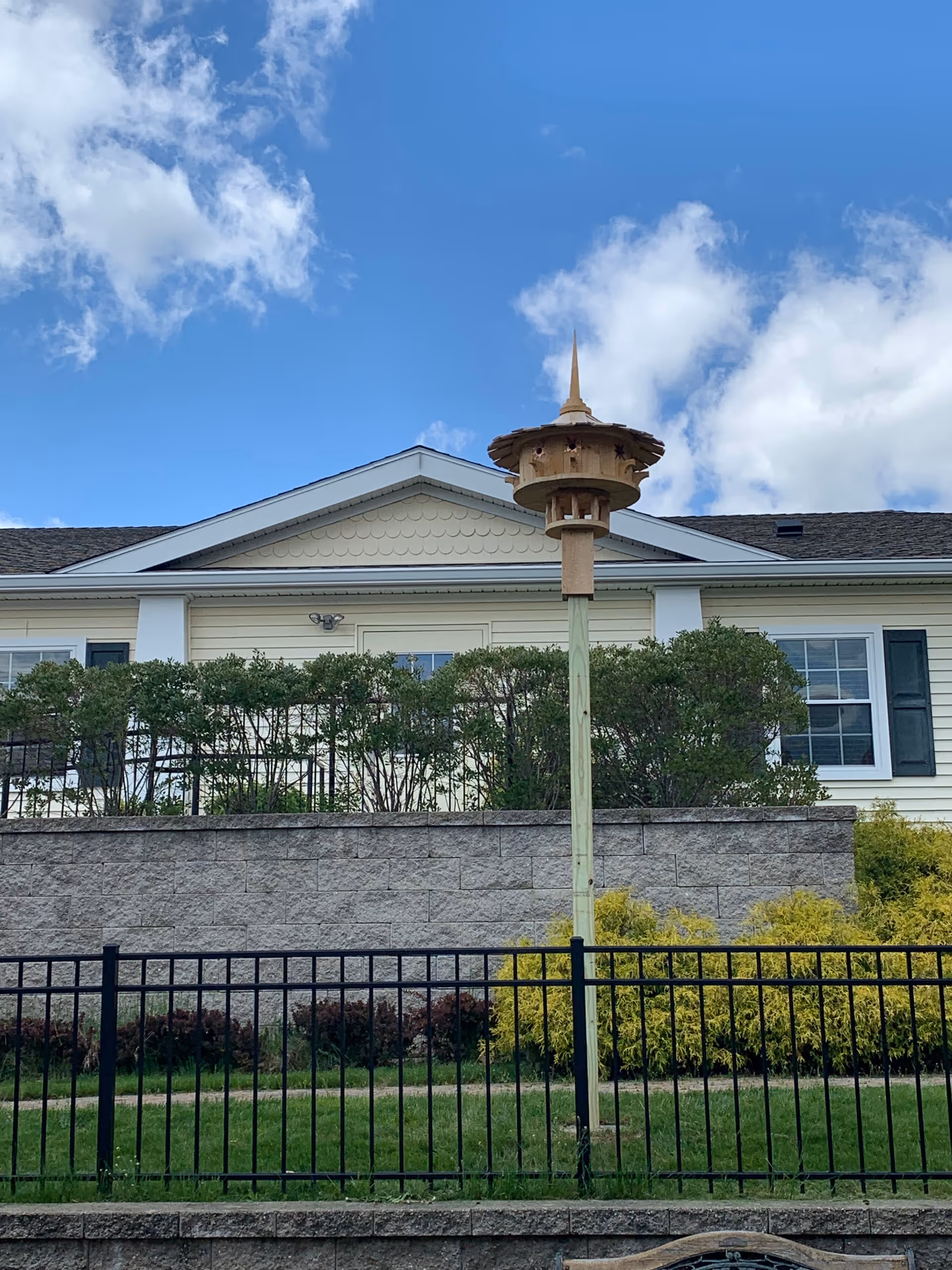A light yellow building with white trim and dark shutters behind a stone retaining wall and black metal fence. In front of the building is a tall wooden birdhouse on a pole. The sky is blue with some white clouds.