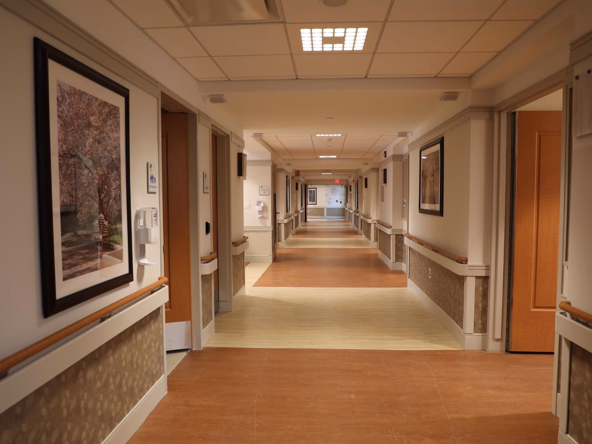 A long, well-lit hallway in a senior living facility with handrails on both sides, framed artwork on the walls, and multiple doorways leading to rooms.