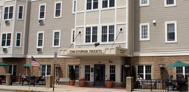 Exterior view of Christopher Heights senior living facility showing the entrance with a sign above the door, outdoor seating areas with people sitting under green umbrellas, American flag, and beige siding with stone accents on the lower part of the building.