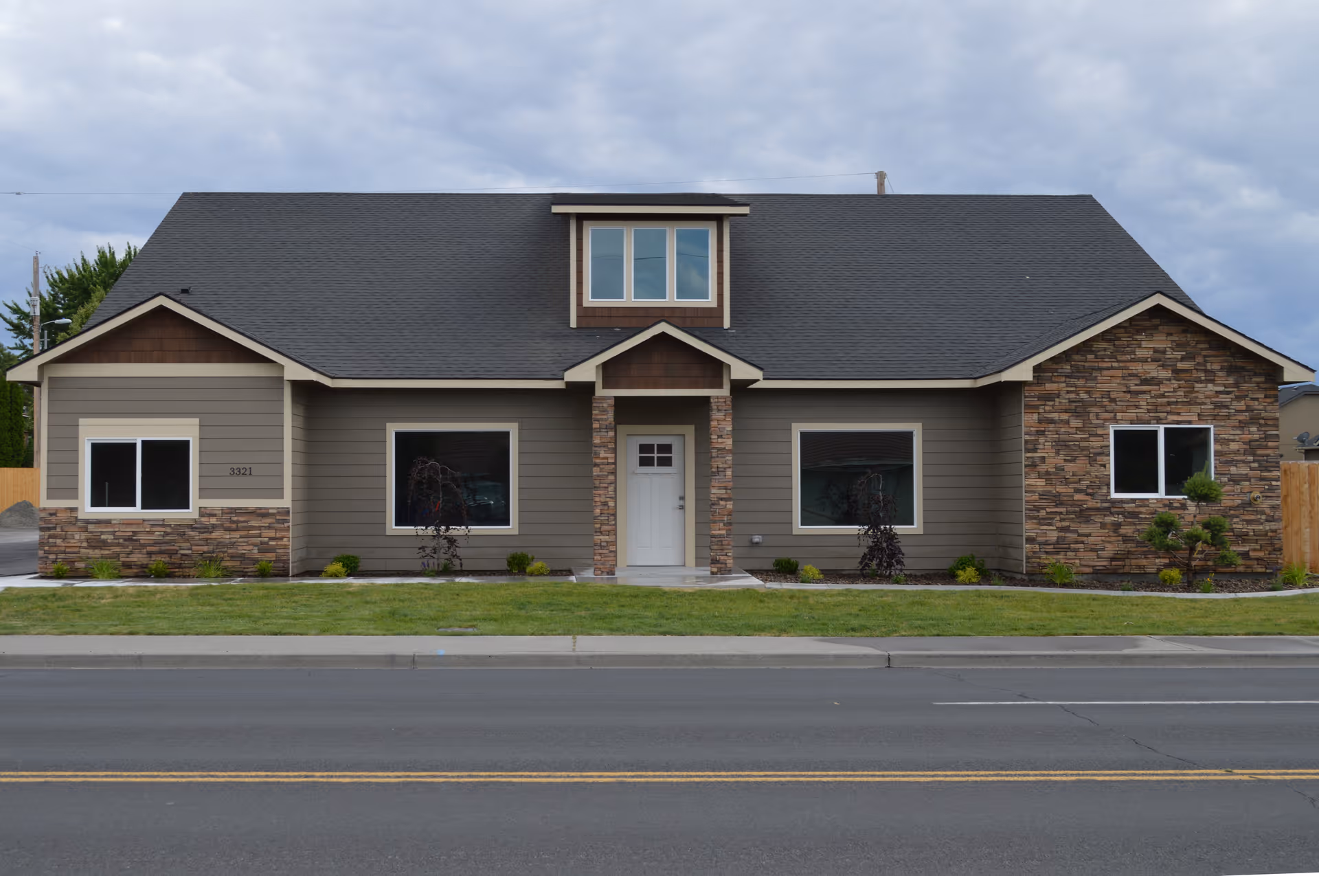 Front exterior view of a single-story building with a dark gray roof, beige and brown siding, stone accents, and a white front door. The building has three large windows and a small dormer window above the entrance. There is a well-maintained lawn and a sidewalk in front of the building under a cloudy sky.