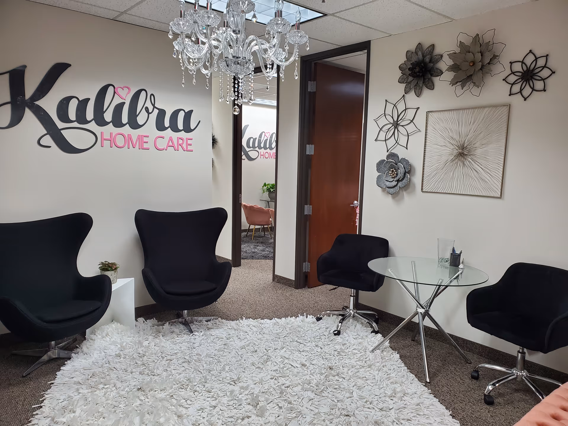 Reception lounge area with Kalibra Home Care sign, chandelier, black chairs, glass table, rug